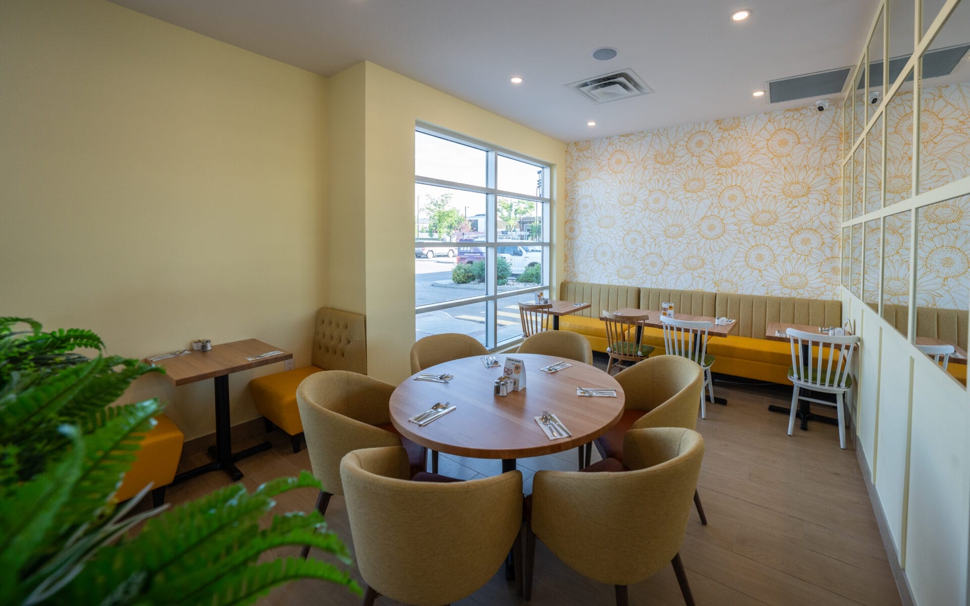 Bright and modern restaurant interior with yellow and beige tones, featuring cushioned chairs around a round wooden table, booth seating along the wall, a large window letting in natural light, and floral-patterned wallpaper.