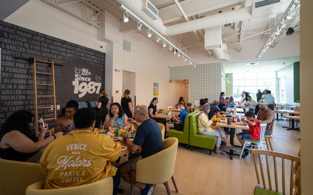 A busy, modern café with people of various ages and backgrounds dining and socializing at wooden tables and green booths under high ceilings with exposed ducts; a wall displays the text "Since 1987" near the entrance.