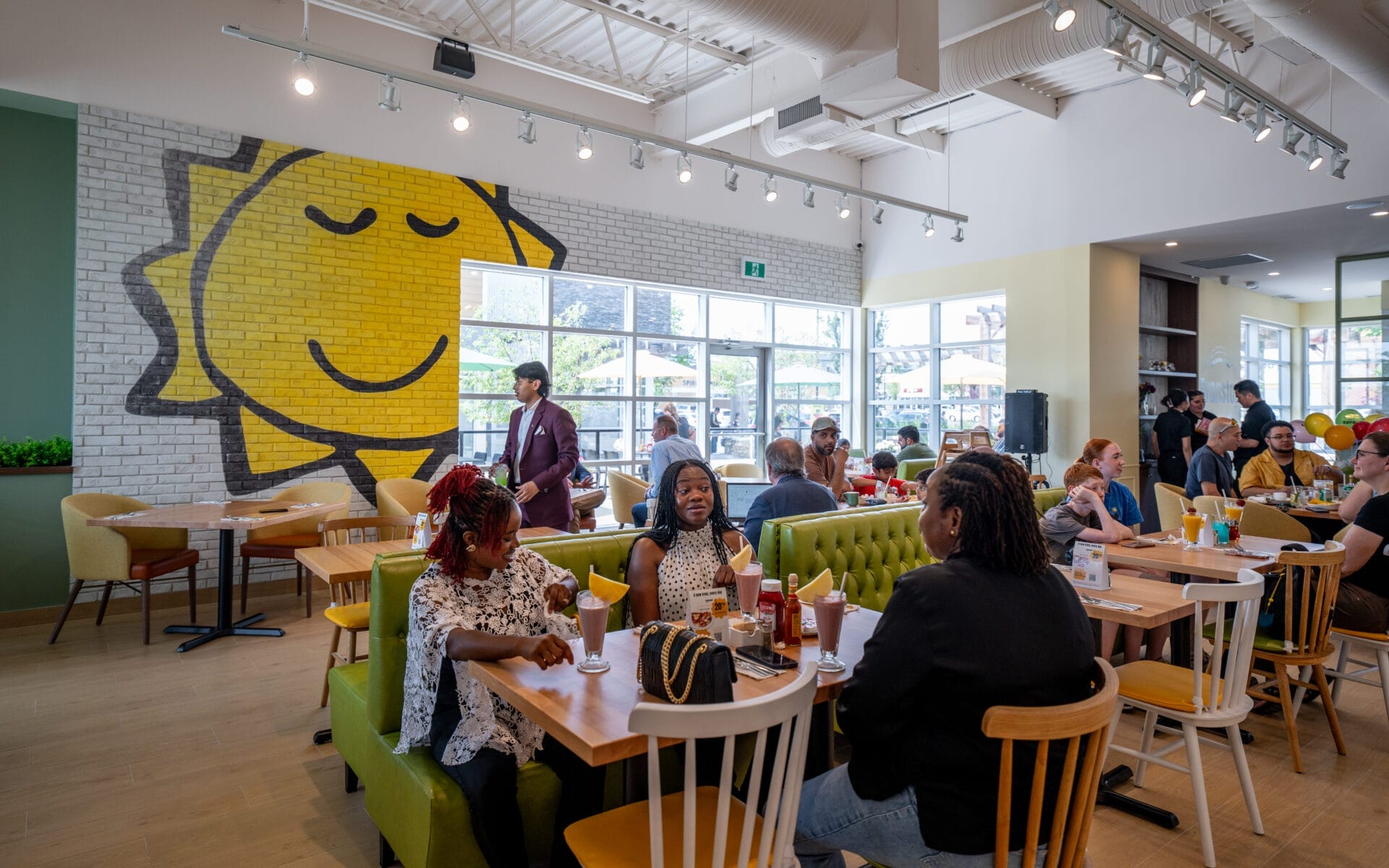 People dining and socializing inside a bright, modern restaurant with large windows and a cheerful yellow sun mural on a brick wall.